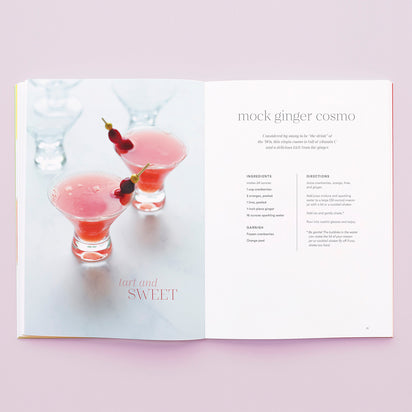 An open "A Collection of Holiday Recipes" book displays two pink mock ginger cosmos—plant-based drinks with red berries on cocktail picks—and a recipe page titled "mock ginger cosmo" with ingredients and directions on a light pink background.