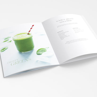 An open "A Collection of Holiday Recipes" book shows a vibrant green smoothie in a jar with a striped straw, highlighting one of many plant-based drinks, with its recipe and directions featured on the opposite page.