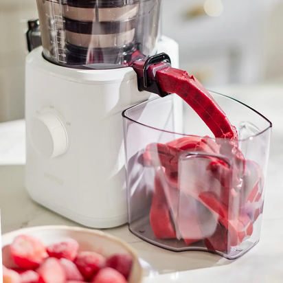 The Sorbet Attachment, a white kitchen accessory, dispenses vibrant red fruit puree into a clear container while a bowl of frozen fruit sits on the brightly lit countertop.