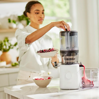 A woman in a bright kitchen uses the Sorbet Attachment on her white juicer, placing berries inside. Fresh juice flows into a container as she prepares ingredients for homemade sorbet on the countertop.