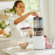 A woman in a bright kitchen uses the Sorbet Attachment on her white juicer, placing berries inside. Fresh juice flows into a container as she prepares ingredients for homemade sorbet on the countertop.