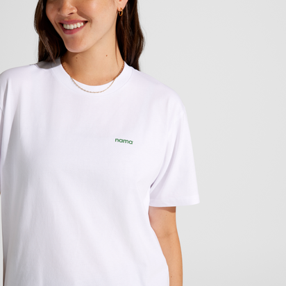 A woman smiles while wearing the Eat More Plants T-shirt, a relaxed-fit white tee with "nama" in small green letters on the chest, standing against a plain light background.