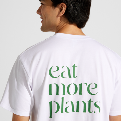 Wearing the Eat More Plants T-shirt, a relaxed fit made from 100% combed cotton, a person smiles and turns to reveal bold green lettering on the back.