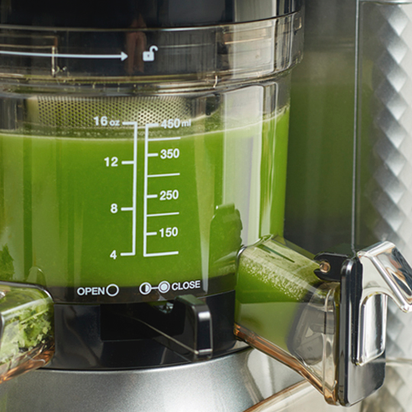 Close-up of the Nama J1 cold press juicer filled with vibrant juices; measurement markings up to 16 oz (450 ml) are visible on the container, with open and close options labeled by the spout.