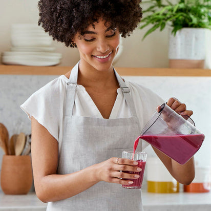 A woman in a light gray apron and white shirt smiles as she pours nutrient-rich juice from the Nama J1 cold press juicer in a bright kitchen, surrounded by plants and wooden utensils.