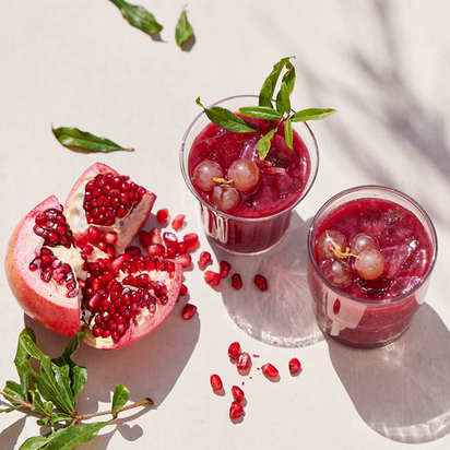 Two glasses of nutrient-rich juice with ice, grapes, and green leaves sit next to a halved pomegranate on a light surface. Sunlight highlights the freshness of juices made with the Nama J1 cold press juicer.