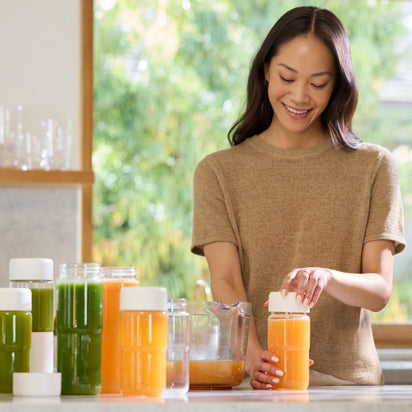 A woman smiles while sealing a Glass Bottle Set, with 17-ounce green and orange juice bottles arranged in front of her on the kitchen counter. Sunlight streams through the window in the background.