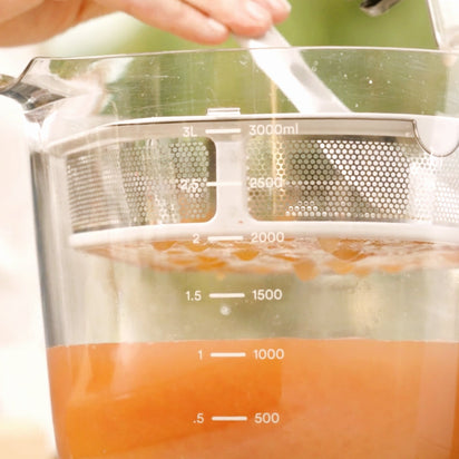 Close-up of the Large Pitcher Set filled with orange liquid to the 1,500 ml mark. It features a custom-fit strainer, measurement markings up to 3,000 ml, and a hand stirring with a spoon—perfect for juice storage.