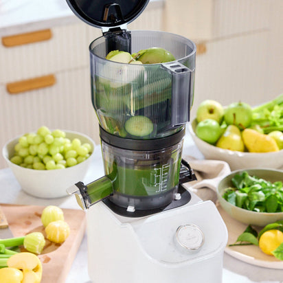 A Large Hopper sits on the counter, filled with chopped green apples and produce. Surrounded by fresh grapes, pears, tomatillos, spinach, and celery, it’s in use for efficient batch juicing of vibrant green juice.