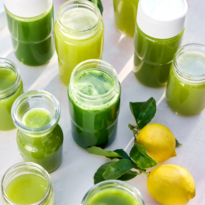 Large Hopper glass jars are filled with green juice from batch juicing and arranged on a white surface beside two fresh lemons with green leaves. Bright natural light highlights the jars’ vibrant colors.