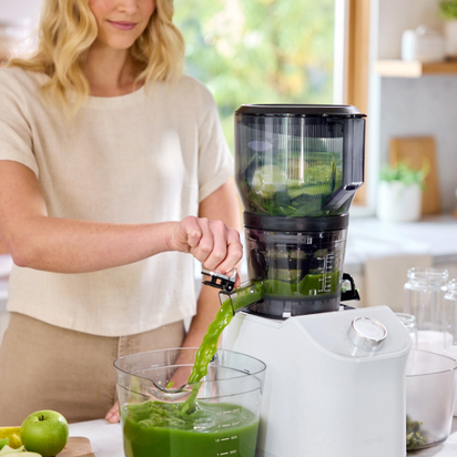 In a bright kitchen, a woman uses the Nama J2 juicer with the Large Hopper to pour fresh green juice into a container, surrounded by green apples and leafy greens—ideal for easy batch juicing.
