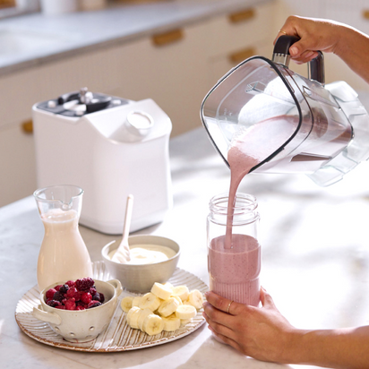 A person pours a pink smoothie into the On-the-Go Glass Bottle with a silicone sleeve. Surrounding ingredients include frozen berries, banana slices, yogurt, and milk—ideal for quick juice blending. A blender base is seen in the background.