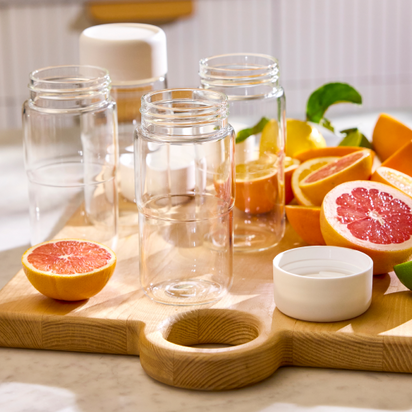 The Glass Bottle Set, featuring three empty bottles and a white lid, sits on a wooden cutting board with grapefruit, orange slices, and lime in a sunlit kitchen—ideal for storing fresh juice.
