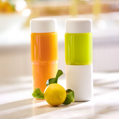 Two On-the-Go Glass Bottles, one with orange liquid and one with green, stand side by side on a white surface. A lemon with green leaves is in front, perfect for enjoying juice anywhere. The background is softly blurred.