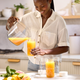 A woman in a light shirt smiles as she pours fresh orange juice from a glass pitcher into a Glass Bottle Set in a bright kitchen, with fruit and mint on the counter.