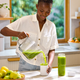 A woman in a white shirt pours green juice from a blender into an On-the-Go Glass Bottle with a silicone sleeve in a sunlit kitchen, surrounded by fruit, preparing to enjoy her fresh drink wherever the day takes her.