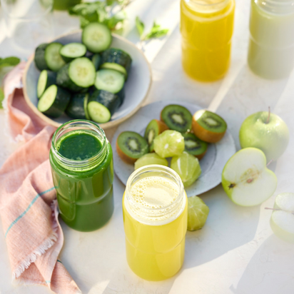 A sunlit table with the Glass Bottle Set filled with green and yellow juice, surrounded by cucumber slices, halved kiwis, lime wedges, and green apples on a pink cloth.