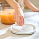 A hand places a white spoon on a white plate atop a marble counter, while a Large Pitcher Set filled with orange drink adds brightness to the scene in the background.