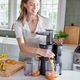 Wearing a light apron, a woman uses the black Nama J1 cold press juicer in a bright kitchen, pouring nutrient-rich juice into a container, with chopped fruit arranged on the counter next to her.